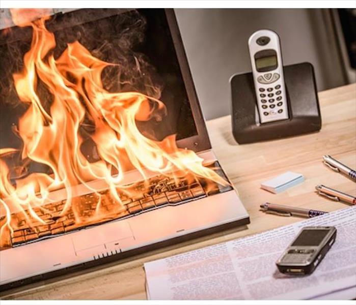 A laptop is sitting on fire on a desk with various other desk items. 